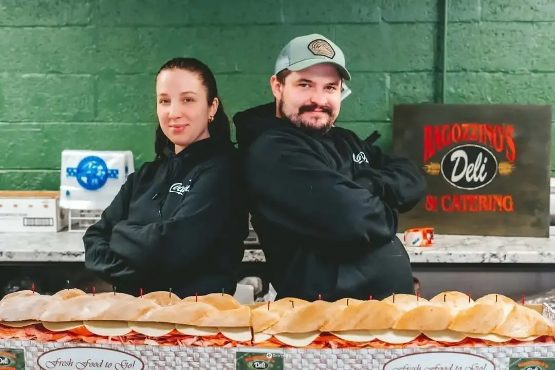 Rob and Margarita, the owners, in black Ragozzino's hoodies behind a party sub in the green-walled deli